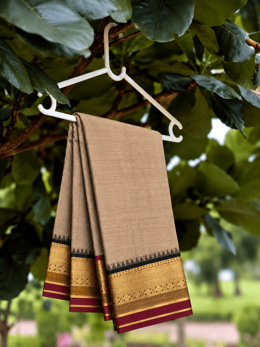 Beige Kanchi cotton saree with a black and gold zari temple border and maroon edge, hung on a hanger under a tree